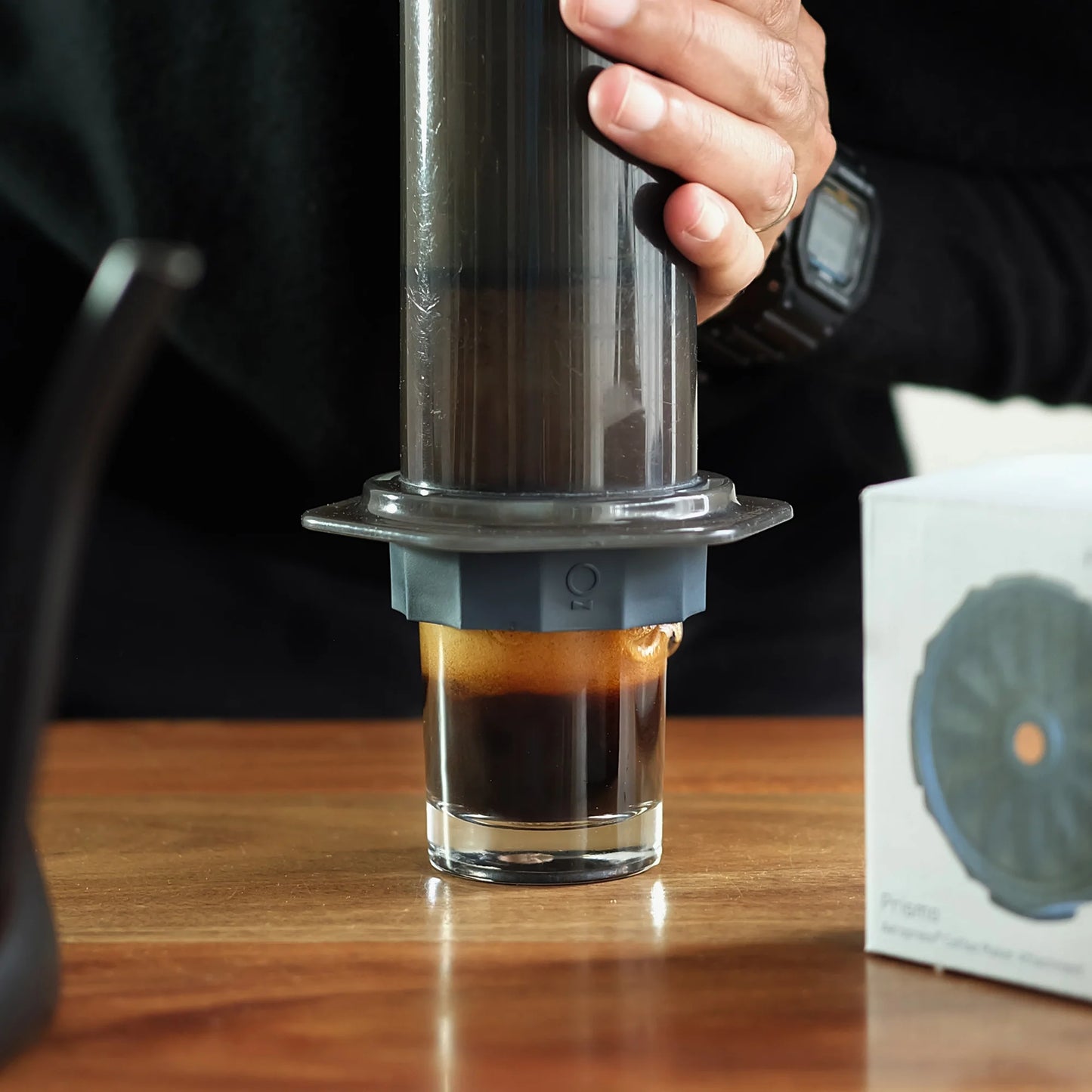 Med et sort ærme og ur brygger en person kaffe i espresso-stil ved hjælp af AeroPress-kaffemaskinen med en Fellow Prismo-tilbehør i et klart glas på et træbord. I nærheden er en kasse med tilbehør til kaffefilter.