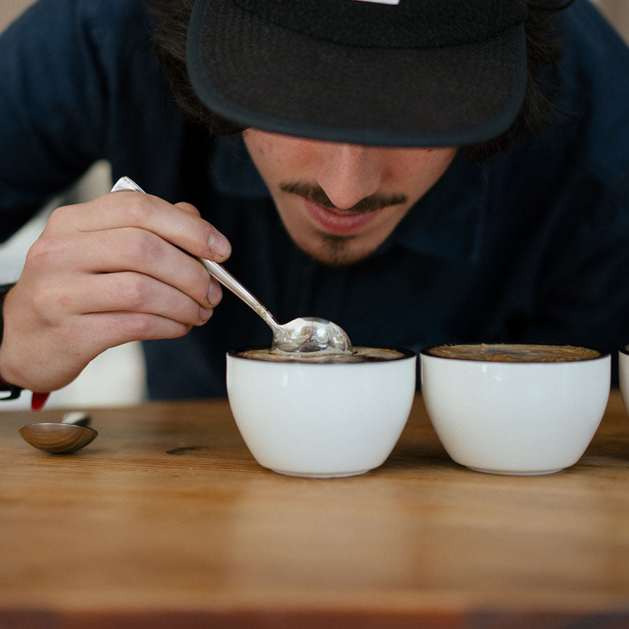 En person i mørk skjorte og kasket prøver kaffe fra et Rhino Coffee Gear, Cupping Skål på et træbord, efter Specialty Coffee Association (SCA) standarder, ved hjælp af en ske.