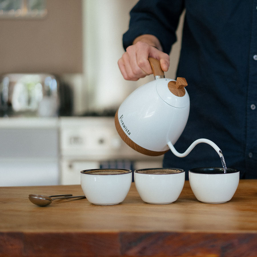 En person hælder varmt vand fra en hvid kedel med en buet tud i tre keramiske kopper på en træbordplade. En ske sidder ved siden af Rhino Coffee Gear Cupping Skål. I baggrunden fuldender et komfur og en brødrister SCA-standard kaffeopsætningen.