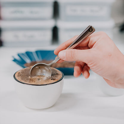 En hånd skummer skum fra en kop kaffe med en ske. Rhino Coffee Gear, Cupping Skål, lavet til keramik perfektion, hviler på en hvid overflade. En sløret baggrund med blå og hvide genstande overholder Specialty Coffee Association (SCA) standarder for entusiasters glæde.