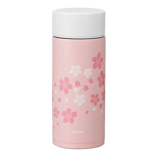 Hario Stick Bottle 350mL Sakura kan prale af kirsebærblomster i hvid og mørk pink på en elegant pink krop med hvidt låg, perfekt til drikke på farten. Mærket "Hario" er elegant printet i bunden.