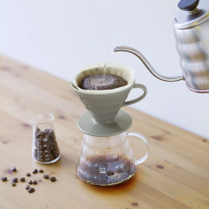 En pour-over kaffe opsætning involverer varmt vand hældt fra en kedel i en Hario Keramisk Drip Tragt med et filter og kaffegrums oven på en glaskaraffel. I nærheden står et bægerglas med kaffebønner og løse bønner på træbordet, alt sammen som en del af den præcise kaffeekstraktionsvidenskab.