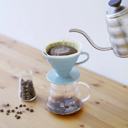 En Hario Keramisk Drip Tragt sidder på en glaskaraffel på et træbord. Vand fra en sølvkedel forstærker kaffeekstraktionen, når den hældes på kaffegrums. I baggrunden fylder friske kaffebønner en lille glasbeholder.