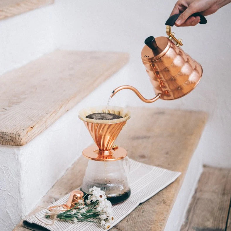 En hånd hælder varmt vand fra en Hario BUONO Cobber-drypkedel i en pour-over kaffemaskine med en glaskaraffel og filterholder på et trætrin. Et stribet klæde med små hvide blomster er ved siden af opsætningen.