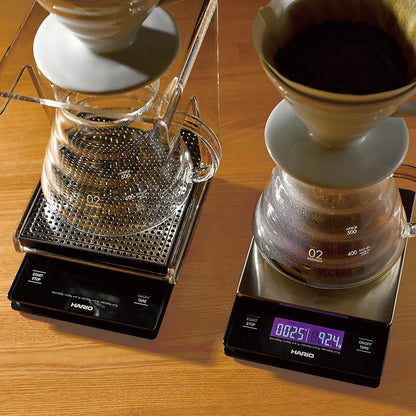 To Hario kaffevægte med digitalt display viser tid og præcise målinger. Den ene holder en glaskaraffel med en V60 Metal Drip Vægt, mens den anden holder en keramisk drypper fyldt med kaffegrums. Begge sidder stilfuldt på træ, ideel til bryggeentusiaster.