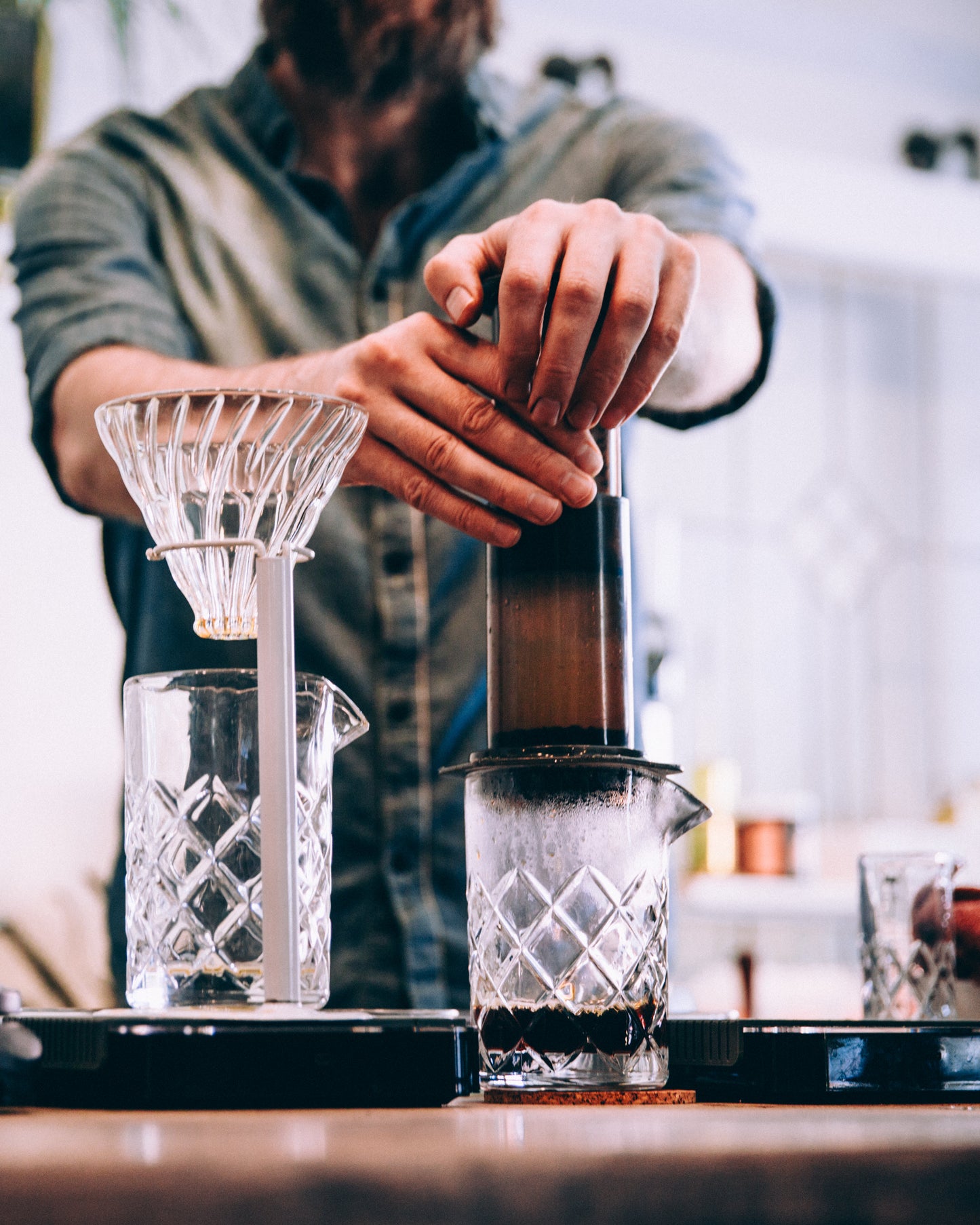 En person i denimskjorte bruger kyndigt en EverBean Aeropress Barista Pack til at brygge kaffe med en kaffevægt i nærheden på trædisken. En glasdryp og et bæger fuldender scenen mod en sløret indendørs baggrund.