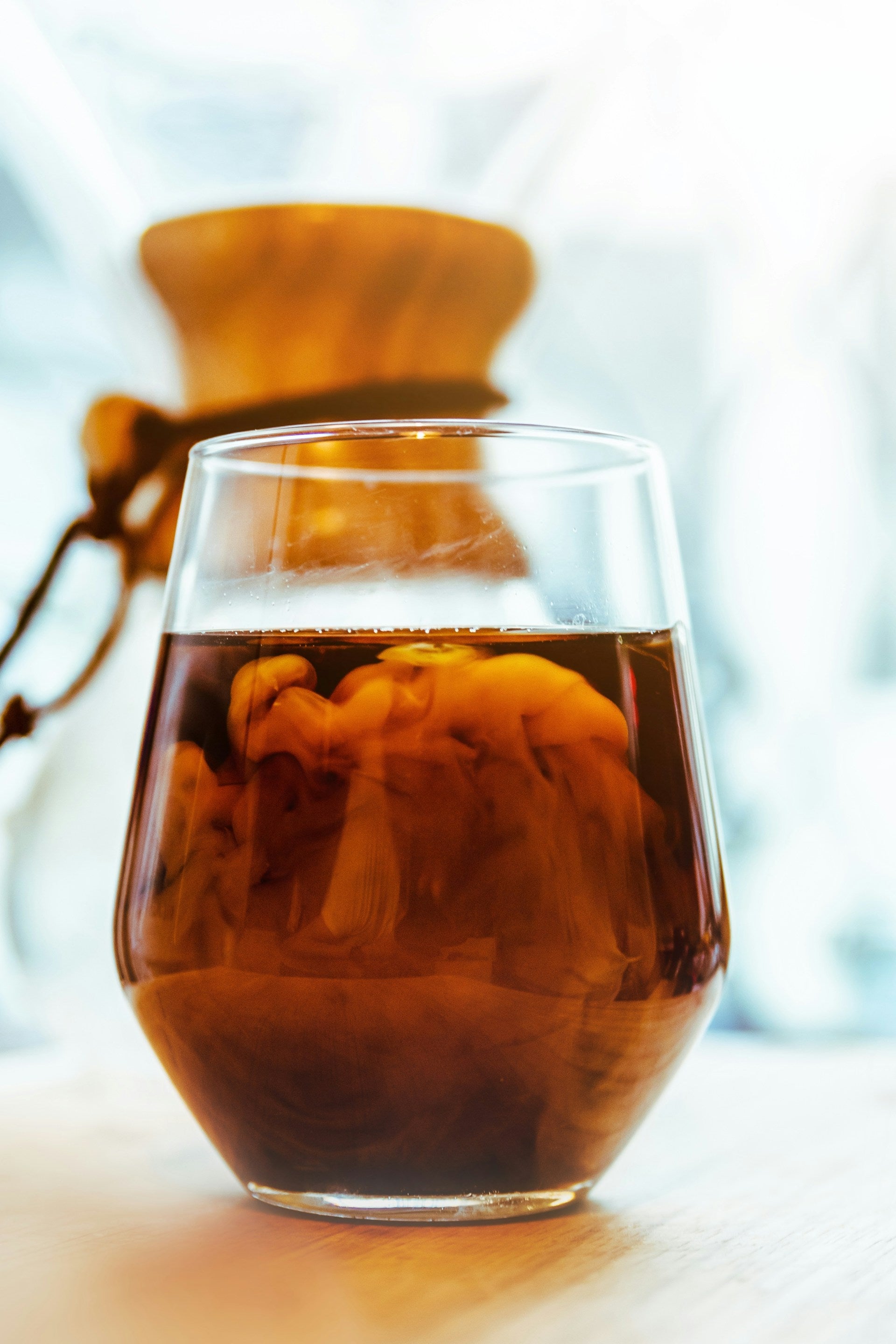 En EverBean ICED Coffee Blend hvirvler med mælk i et glas på en lys træoverflade. I baggrunden fremhæver en sløret Chemex den glatte rigdom af 100% Arabica-bønne-koldbryg. Scenen er smukt oplyst af naturligt lys.