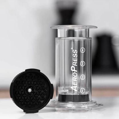 Et nærbillede af en AeroPress med stemplet fjernet afslører det gennemsigtige nummererede kammer, opretstående på en hvid overflade. Det sorte stempel, der har et sekskantet design, antyder espresso-lignende crema, mens en sløret baggrund tilføjer dybde. Produkt: AeroPress med Flow Control Filter Cap fra Aeropress.