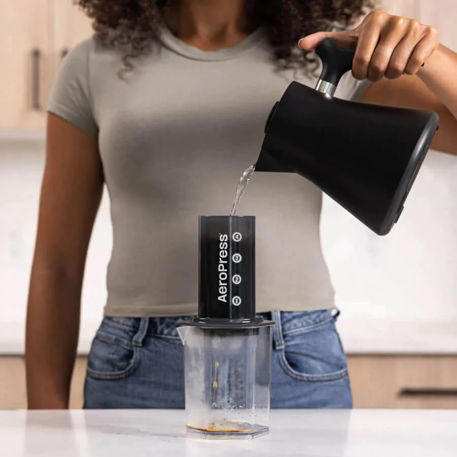 En person i en grå t-shirt hælder varmt vand fra en sort kedel i en Aeropress kaffemaskine, placeret over en Aeropress Karaffel. Køkkenet danner med sine lyse skabe den perfekte baggrund for kaffe brygning.