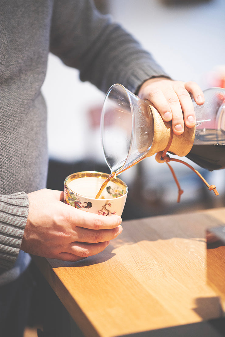 En person i en grå sweater hælder forsigtigt glat Kenya-kaffe fra en Chemex Barista Pakke (6 pax) fra Chemex & EverBean i en blomster-mønstret kop på en træoverflade, med varm belysning, der fremhæver hænderne og en kunstfærdig skænkning.